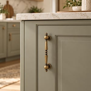 May include: A vertical cabinet handle with a distressed antique brass finish, featuring a central cylindrical bar with decorative rounded accents at the top, bottom, and middle, mounted on a sage green cabinet door with a recessed panel design.