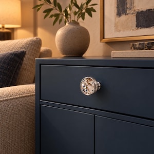 Op de afbeelding: Een ronde meubelknop met een zichtbaar mechanisch uurwerk met metalen tandwielen, kleine schroeven en blauwe accenten in een gepolijst zilverkleurig frame, gemonteerd op een donkerblauwe ladefront met een gladde, matte afwerking.
