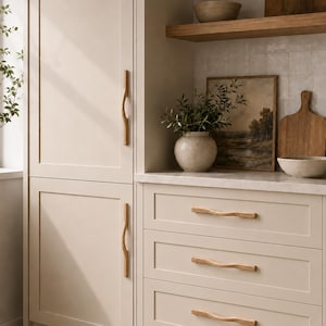 May include: A set of light-coloured wooden cabinet handles with a wavy, organic shape installed on cream-coloured kitchen cabinetry. The hardware features a smooth, natural wood grain finish and is mounted vertically on doors and horizontally on drawers.