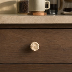 May include: A circular cabinet knob with a rough, textured, light beige stone-like surface attached to a dark brown wooden drawer. The knob features a natural, uneven finish with visible pores and crevices, contrasting against the smooth wood grain.