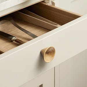 May include: A light beige kitchen drawer featuring a cone-shaped wooden knob with a visible grain pattern. The drawer is partially open, revealing a light wood interior organizer containing a metal spatula and a knife with a wooden handle.