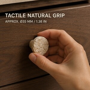May include: A round, beige, textured stone cabinet knob with a rough, porous surface. The text TACTILE NATURAL GRIP and APPROX. Ø35 MM / 1.38 IN is visible above the knob. The knob is mounted on a dark brown wood drawer front.