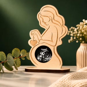 May include: A light-coloured wooden cutout sculpture depicting the silhouette of a pregnant person. The figure holds a small wooden heart, and a circular black and white ultrasound image is embedded in the centre of the belly area on a base.