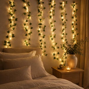 Op de afbeelding: Een reeks verticale lichtsnoeren verweven met kunstmatige groene klimopbladeren die tegen een muur hangen. De warmwitte LED-lampjes zijn verdeeld over de ranken, wat een gloeiend effect creëert. Een keramische vaas met groen staat in de buurt.