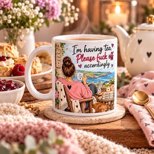 Peut inclure: Un mug en céramique blanche illustré d'une personne aux cheveux en chignon assise sur une chaise en bois face à un paysage côtier. Le texte indique, I'm having tea. Please fuck off accordingly. Un petit chien est assis près de la chaise.