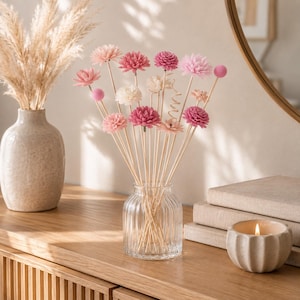 May include: A collection of decorative floral stems featuring various shades of pink and cream-colored blossoms attached to thin wooden sticks. These stems are arranged inside a clear, vertically ribbed glass vase, creating a bouquet display.