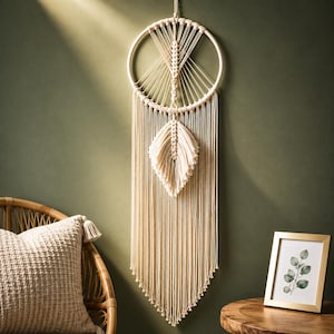 Puede incluir: Un tapiz de macramé color crema con un aro de madera circular, patrones de nudos intrincados y un elemento central en forma de hoja. Largos flecos verticales cuelgan del aro, creando un diseño simétrico que termina en punta.