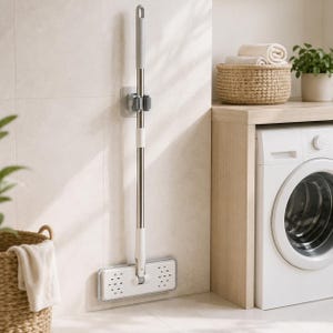 Peut inclure: Un balai à plat avec un manche en métal argenté et une tête rectangulaire blanche dotée d'une grille de petits trous d'aération. Le balai est maintenu en position verticale contre un mur par une attache murale en plastique gris.