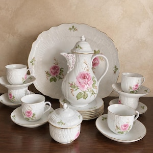 Peut inclure: Un service à thé en porcelaine blanche orné d'un motif de roses roses, de feuilles vertes et d'un liseré doré. L'ensemble comprend une théière avec couvercle, un sucrier avec couvercle, des tasses avec soucoupes et un grand plat de service.