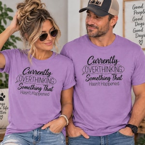 Pode incluir: Duas t-shirts de manga curta a condizer em violeta claro com o texto preto Currently OVERTHINKING Something That Hasn't Happened. As t-shirts têm um decote redondo e um corte padrão com o mesmo design gráfico na parte da frente.