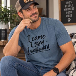 May include: A heathered blue short-sleeve t-shirt featuring black text that reads I came I saw I overthought it. The shirt is paired with blue denim jeans and a black and tan baseball cap with a mountain graphic on the front patch.