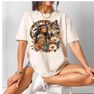 May include: A cream-colored short-sleeve t-shirt featuring a graphic print of a person wearing a feathered headband and patterned clothing, positioned next to a wolf. The design includes a circular wooden frame, colorful flowers, and feathers.