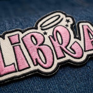 May include: An embroidered patch featuring the word LIBRA in pink thread with a thick black outline and a white background. The design includes a small halo above the letter I and two tiny stars, all stitched onto a dark blue denim fabric surface.