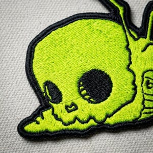 May include: An embroidered patch featuring a bright lime green skull design with bold black outlines and deep black eye sockets. The skull has a rounded shape with a jagged jawline and is rendered in a dense, textured thread stitch pattern.