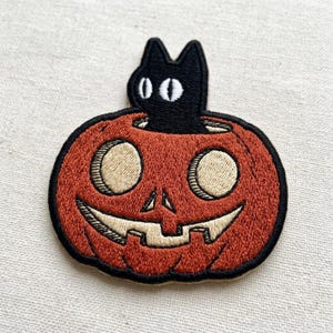 May include: An embroidered patch featuring a black cat silhouette emerging from the top of a round orange pumpkin. The pumpkin has a carved face with light beige eyes and a wide, toothy grin, all outlined in thick black thread stitching.