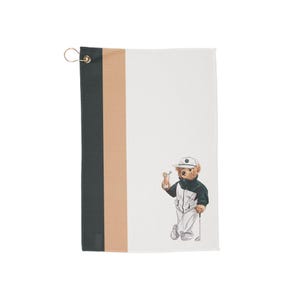 May include: A rectangular golf towel featuring a vertical color-blocked design in black, tan, and white. The white section displays a graphic of a teddy bear wearing a white cap and a black and white jacket, holding a drink and a golf club.