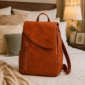May include: A cognac-coloured faux leather backpack featuring a large top flap, a vertical zippered front pocket with gold-toned hardware, a top handle, and adjustable shoulder straps. The design includes a diagonal seam across the front panel.