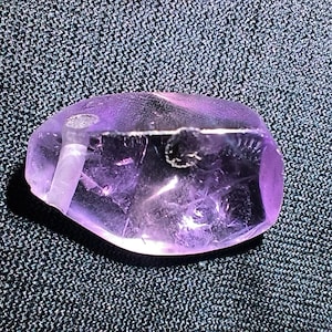 May include: A translucent, irregular, purple amethyst gemstone with a polished surface. The stone features visible internal inclusions, natural fractures, and a small, circular drilled hole near the left edge, showcasing its raw, crystalline structure.