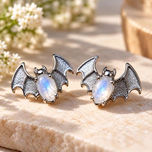 May include: A pair of stud earrings shaped like bats with dark metallic wings and an oval-shaped iridescent moonstone cabochon set in the center of each body. The wings feature a textured surface, and the stones display a soft blue glow.