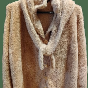 Op de afbeelding: Een beige, langharig vest met lange mouwen en een zachte, getextureerde pool. Het kledingstuk heeft een brede, geïntegreerde sjaalachtige kraag die aan de voorzijde naar beneden valt, vastgezet met vier ronde, lichtgekleurde knopen in een rij.