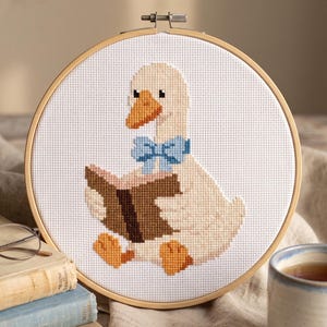 May include: A circular wooden embroidery hoop containing a cross-stitch pattern of a light cream-colored goose with an orange beak and feet. The goose wears a light blue bow tie and holds an open brown book on a white Aida fabric background.