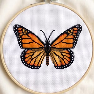 May include: A circular wooden embroidery hoop containing white fabric with a cross-stitch design of a monarch butterfly. The butterfly features vibrant orange wings with black veins and borders, centered on the fabric within the light wood frame.