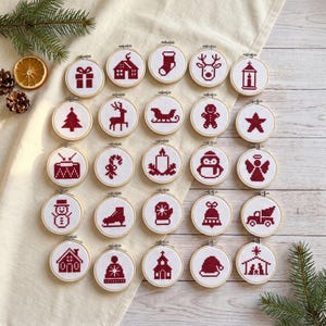 May include: A collection of twenty-five small circular wooden embroidery hoops, each featuring a unique red cross-stitch design on white fabric. The motifs include Christmas icons like a gift, house, stocking, reindeer, tree, sleigh, gingerbread man, star, drum, candy cane, candle, penguin, angel, snowman, skate, mitten, bell, truck, hat, church, and nativity scene.