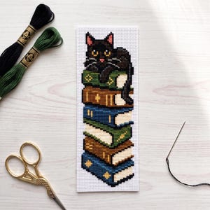 May include: A rectangular cross-stitch bookmark on white Aida fabric featuring a black cat with pink-tipped ears resting atop a vertical stack of six colorful books in shades of green, brown, and blue, accompanied by embroidery floss and scissors.