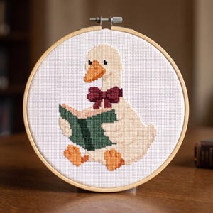 May include: A circular wooden embroidery hoop containing a cross-stitch pattern on white fabric. The design features a light cream-coloured goose with an orange beak and feet, wearing a dark red bow tie while holding an open dark green book.