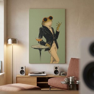 May include: A wall art print featuring a stylized illustration of a frog wearing orange-rimmed sunglasses and a black suit jacket. The frog stands beside a small pedestal table holding a cocktail glass against a solid sage green background.