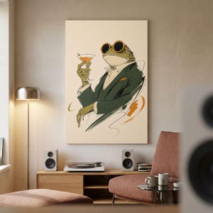 May include: A wall art print featuring an illustration of a frog wearing round orange-rimmed sunglasses and a dark green suit jacket. The frog is holding a cocktail glass with an orange liquid, rendered in a sketch-like style on a cream background.