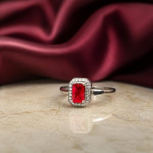 Puede incluir: Un anillo de tono plateado con una piedra central rectangular de color rojo vibrante. La piedra está rodeada por un halo de pequeñas piedras claras y brillantes engastadas en el marco de metal. El aro es liso, pulido y refleja la luz.