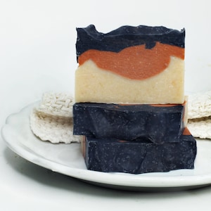 May include: Three handmade soap bars stacked on a white plate. The top bar has layers of black, orange, and cream colors. The bottom two bars are dark with orange accents. Two white, textured scrubbies are on the plate.
