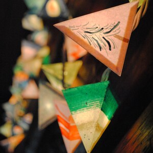 May include: A string of triangular paper lanterns with various colors and patterns, including green, pink, and white. The lanterns are decorated with black brushstrokes.