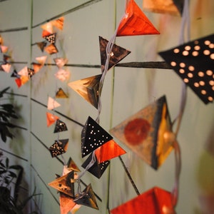 May include: A string of triangle-shaped paper lanterns with different patterns and colors, including black, red, orange, and white. The lanterns are strung together with white wire and are lit up with small white lights.