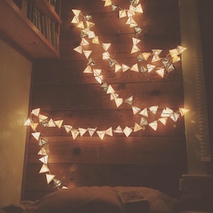 May include: A string of triangle-shaped paper lanterns with warm white lights, hanging on a wood wall. The lanterns are made of white paper with a gold glitter finish.