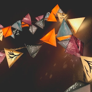 May include: A string of paper lanterns in various colors, including orange, blue, purple, and white, with a white string running through them. The lanterns are shaped like triangles and have a variety of patterns, including dots, stars, and branches.