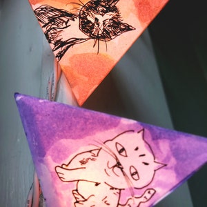 May include: A string of triangle-shaped paper lanterns with hand-drawn images of cats. The lanterns are lit from inside, creating a warm glow.