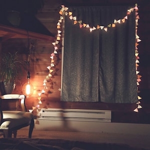 May include: A string of colorful paper triangle lights hangs in front of a window with gray curtains. The lights are lit and cast a warm glow on the room. A light bulb hangs from the ceiling above a chair.
