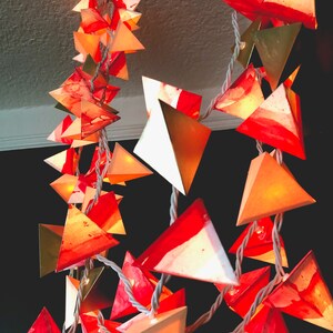 May include: A string of paper lanterns with a white cord. The lanterns are made of white paper with red and gold accents. The lanterns are shaped like triangles and are lit from within.