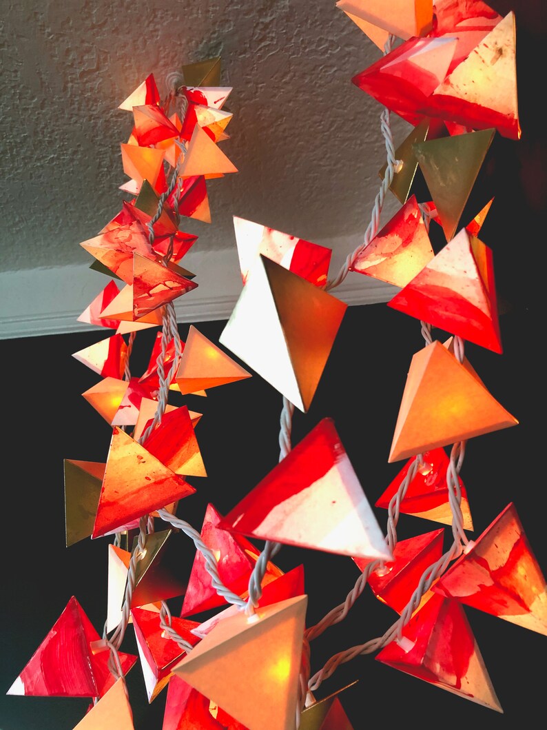 May include: A string of paper lanterns with a white cord. The lanterns are made of white paper with red and gold accents. The lanterns are shaped like triangles and are lit from within.