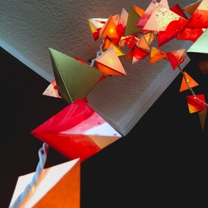 May include: A string of paper lanterns in various shades of red, orange, and gold, with a few green lanterns, hanging from a ceiling. The lanterns are illuminated from within, creating a warm and festive atmosphere.
