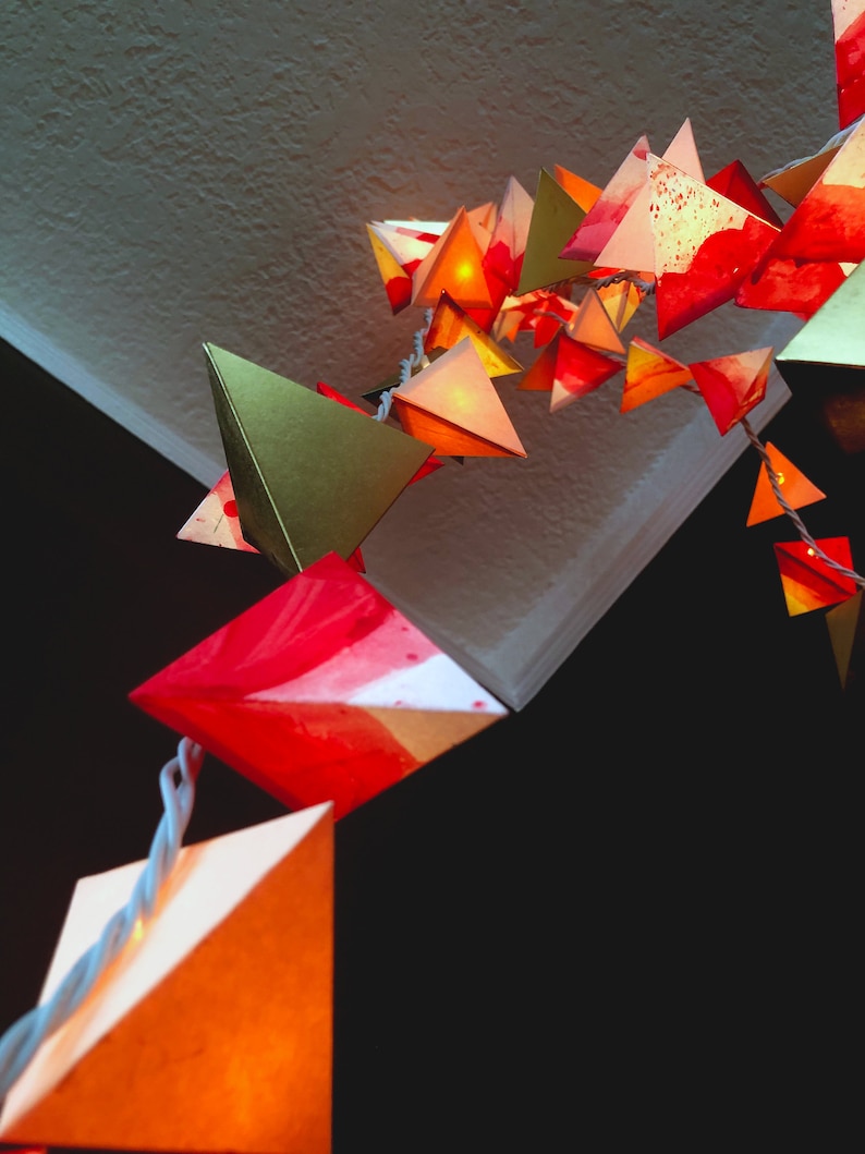 May include: A string of paper lanterns in various shades of red, orange, and gold, with a few green lanterns, hanging from a ceiling. The lanterns are illuminated from within, creating a warm and festive atmosphere.