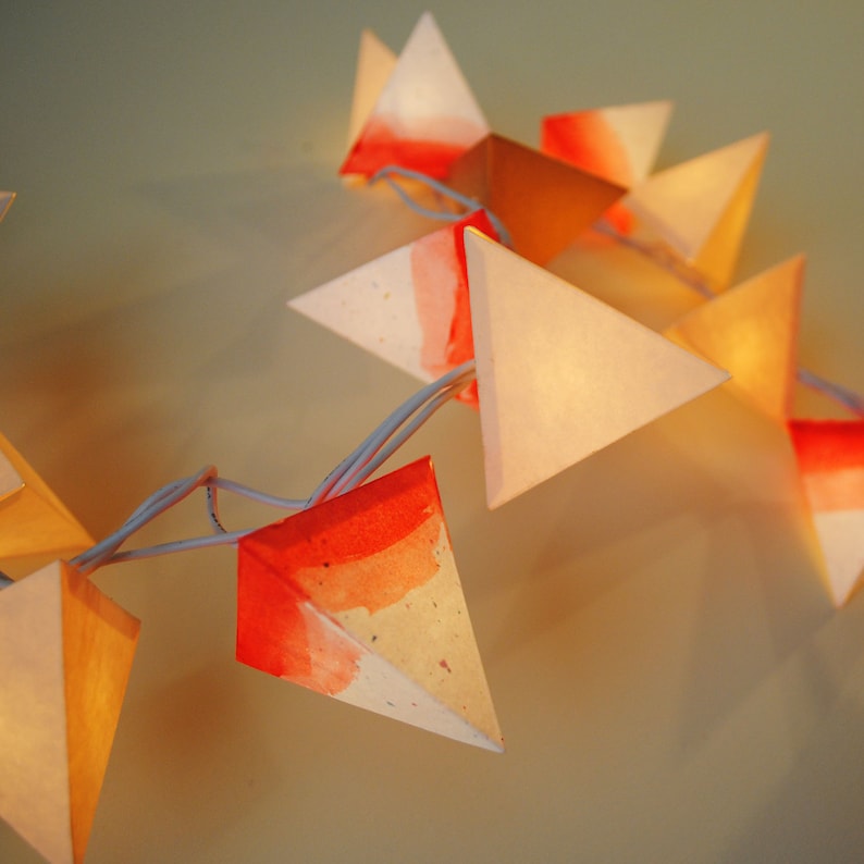 May include: A string of paper lanterns with a white cord. The lanterns are triangular and made of white paper with a red and orange watercolor design. The lanterns are lit from inside, creating a warm glow.