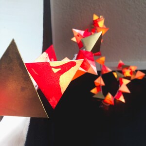 May include: A string of red and gold origami star lights. The stars are strung together with clear wire and have a warm glow.