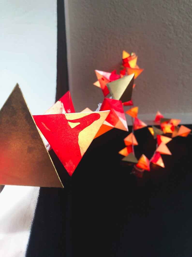 May include: A string of red and gold origami star lights. The stars are strung together with clear wire and have a warm glow.
