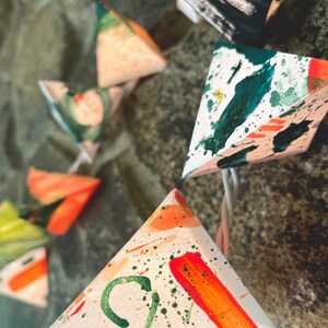 May include: A string of triangle-shaped lights with a white background and colorful abstract designs. The lights are painted with green, orange, and red accents.