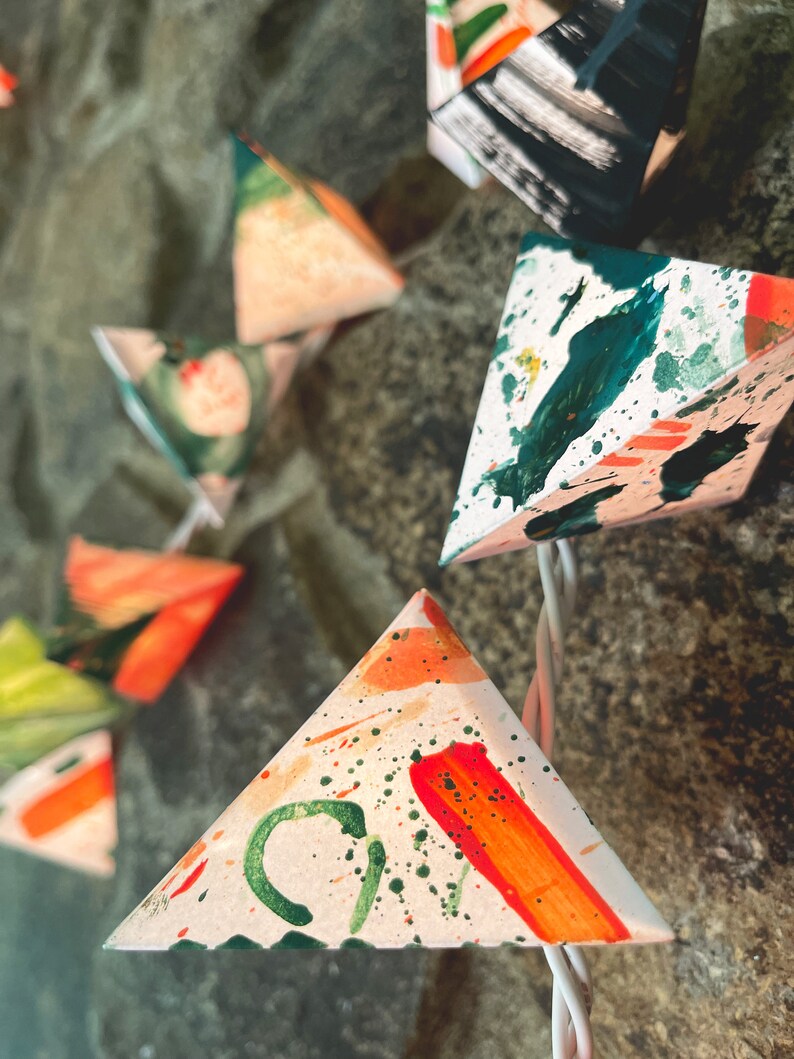 May include: A string of triangle-shaped lights with a white background and colorful abstract designs. The lights are painted with green, orange, and red accents.