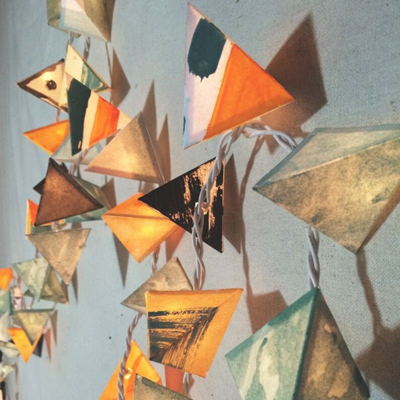 May include: A string of triangle-shaped paper lanterns with a variety of abstract patterns and colors, including orange, yellow, black, brown, and green. The lanterns are strung together with white wire and are lit from within, creating a warm and inviting glow.