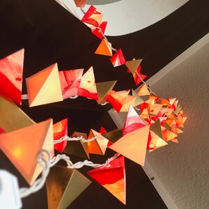 May include: A string of lights with red and gold triangular paper lanterns. The lanterns are lit and glowing, creating a warm and festive atmosphere.
